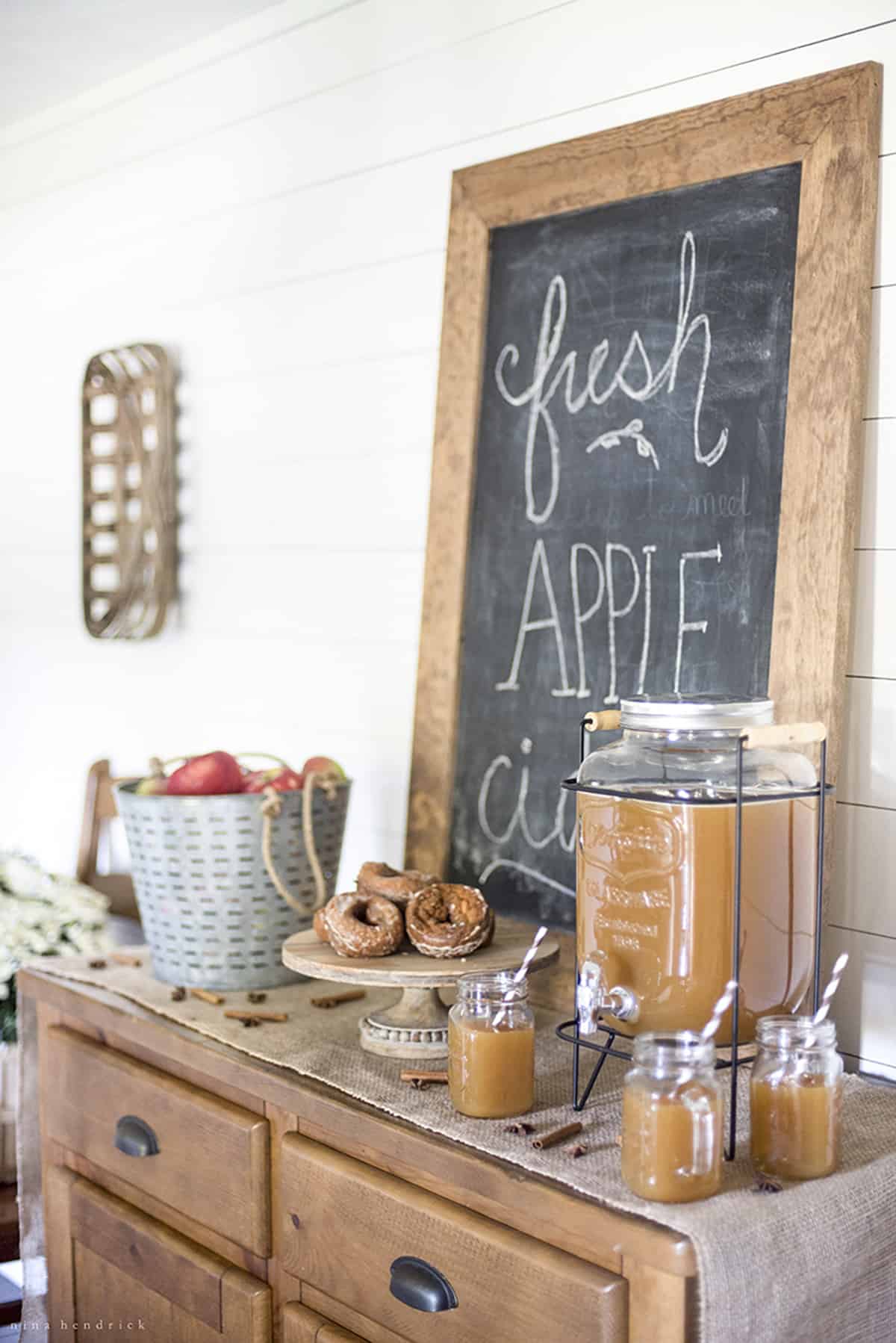An Apple Cider Bar on a wooden stand with burlap tablecloth, beverage dispenser, plate of donuts, and a chalkboard that says "Fresh Apple Cider".