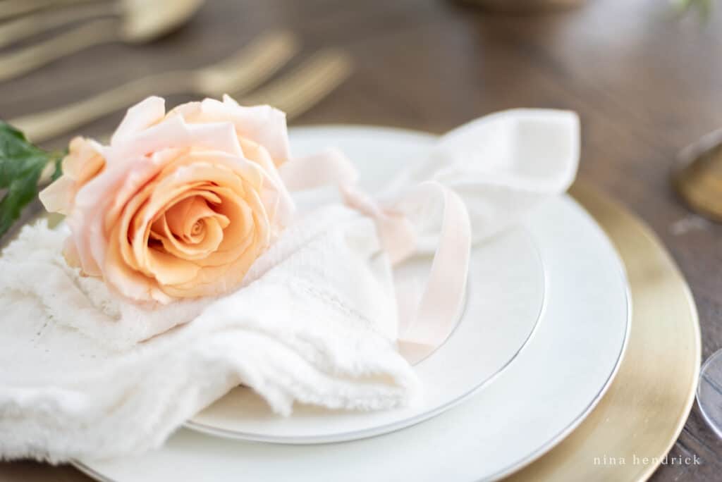 Place setting with blush rose and white frayed linen napkin, white plates, and a gold charger. 