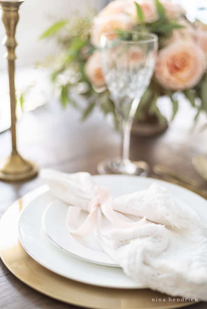 White frayed linen napkin with a blush ribbon over white plates on a gold charger. 