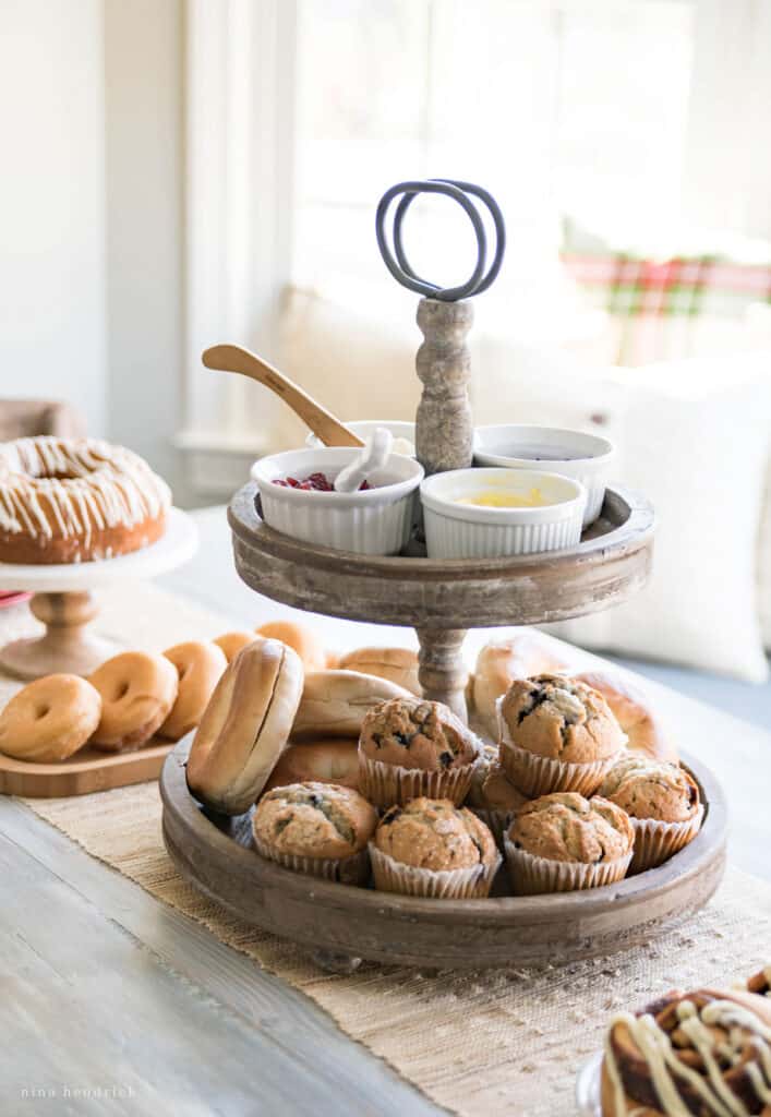Two-tiered wooden tray with muffins and bagels with toppings and donuts and cake in background