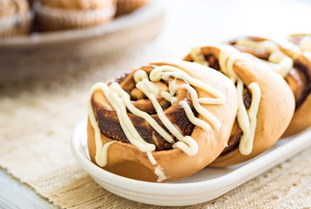 Cinnamon buns with icing on a white tray with a burlap runner below