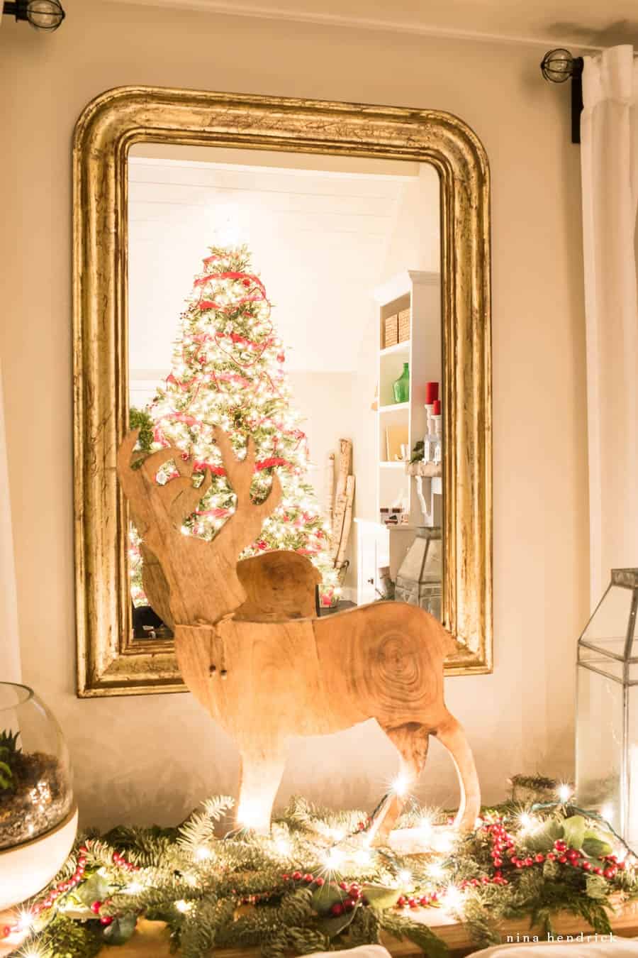 Christmas tree reflected in mirror with wooden deer statue and evergreen garland with Christmas lights