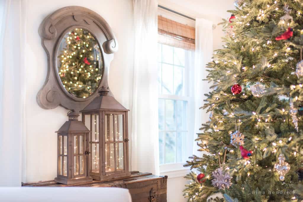 Gray rustic lanterns with twinkle lights in front of a gray curved mirror beside a Christmas tree with red and white ornaments