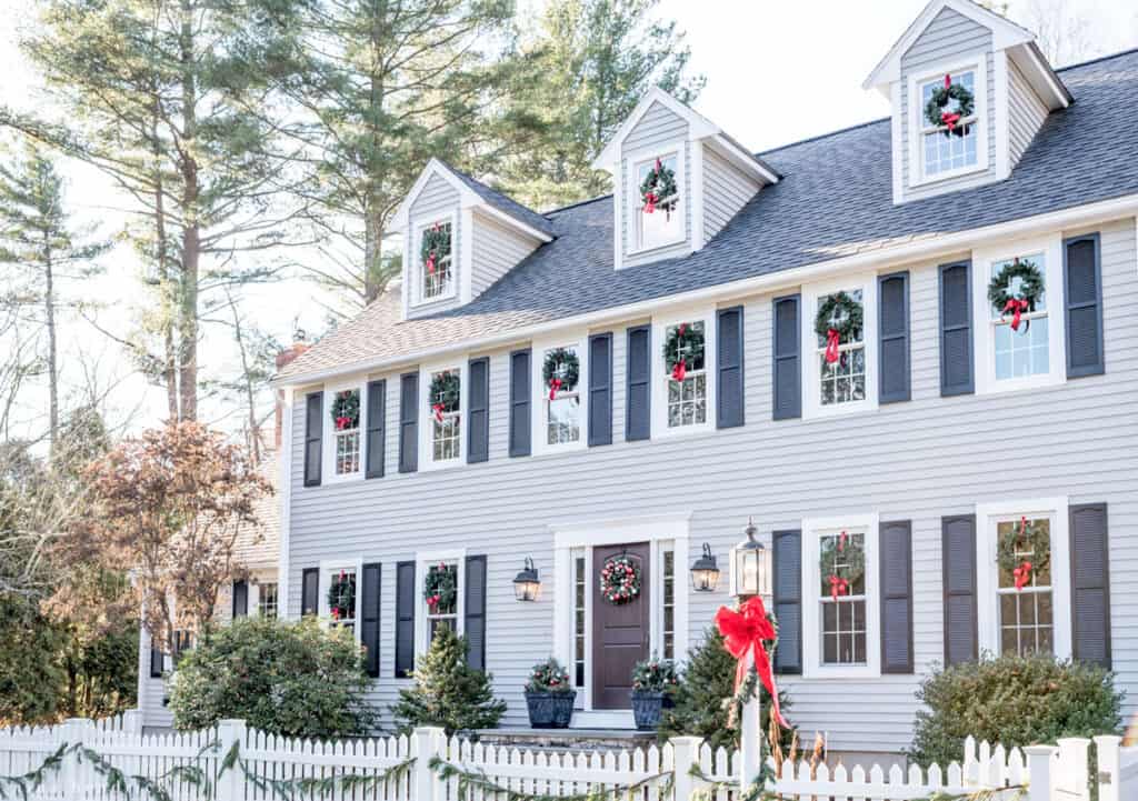 Exterior of a gray colonial home with classic outdoor Christmas decorations like wreaths in each window and red ribbons. 