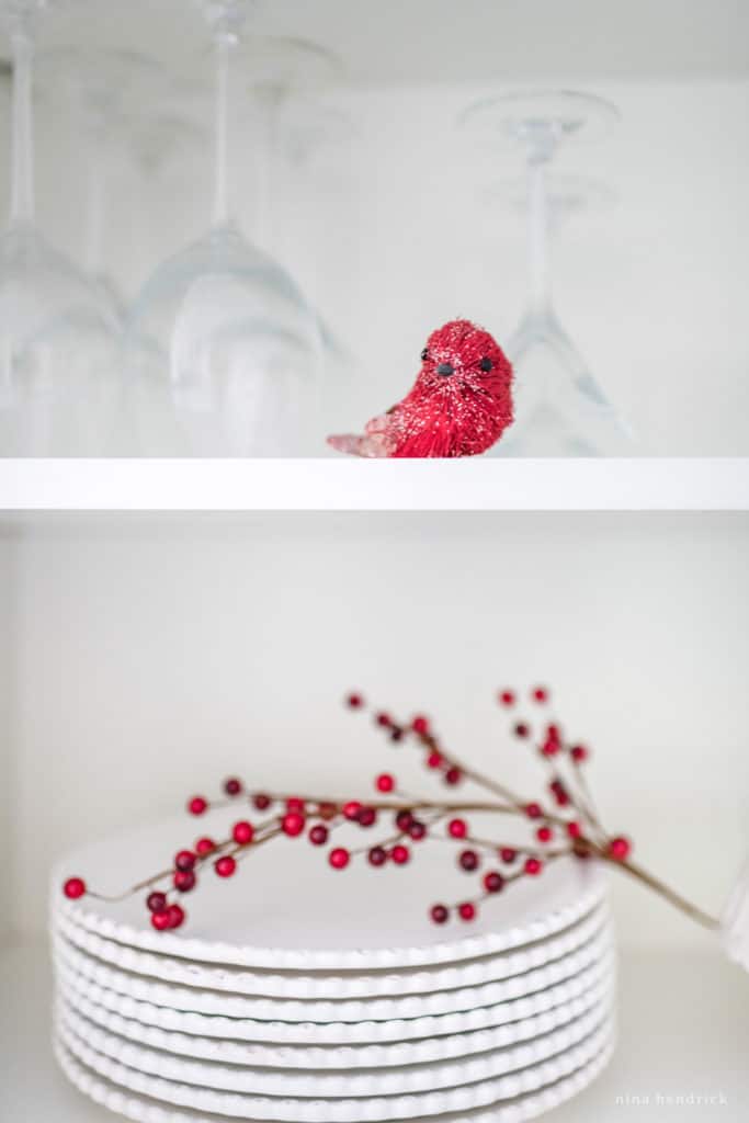 Red bird and berries on a white kitchen shelf with white plates. 