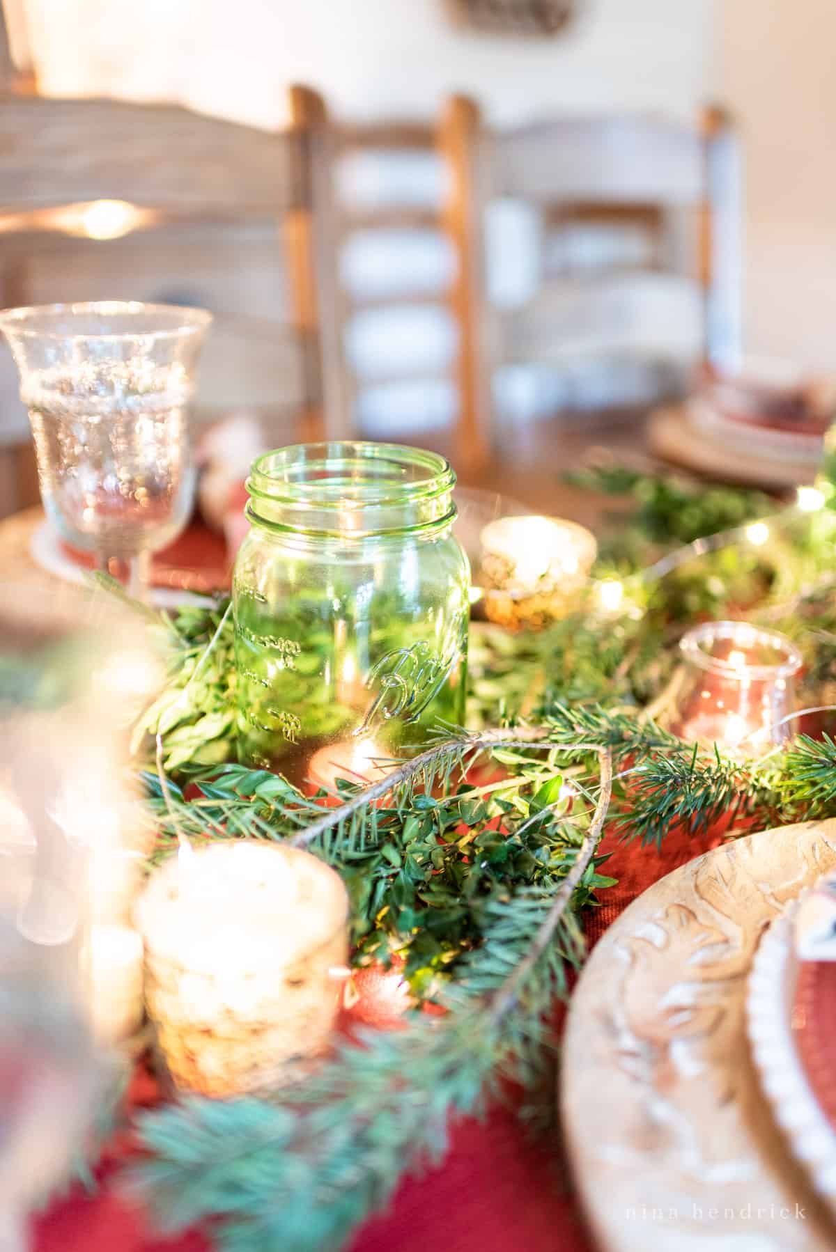 Gather inspiration for your holiday entertaining with this Classic Christmas Tablescape featuring traditional reds and greens. Green mason jar in evergreen centerpiece with holiday twinkle lights and candles