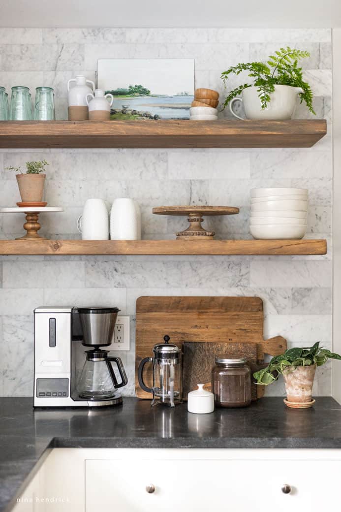 Warm wood floating shelves with plants and decor over a white cabinet and dark countertop in a kitchen makeover