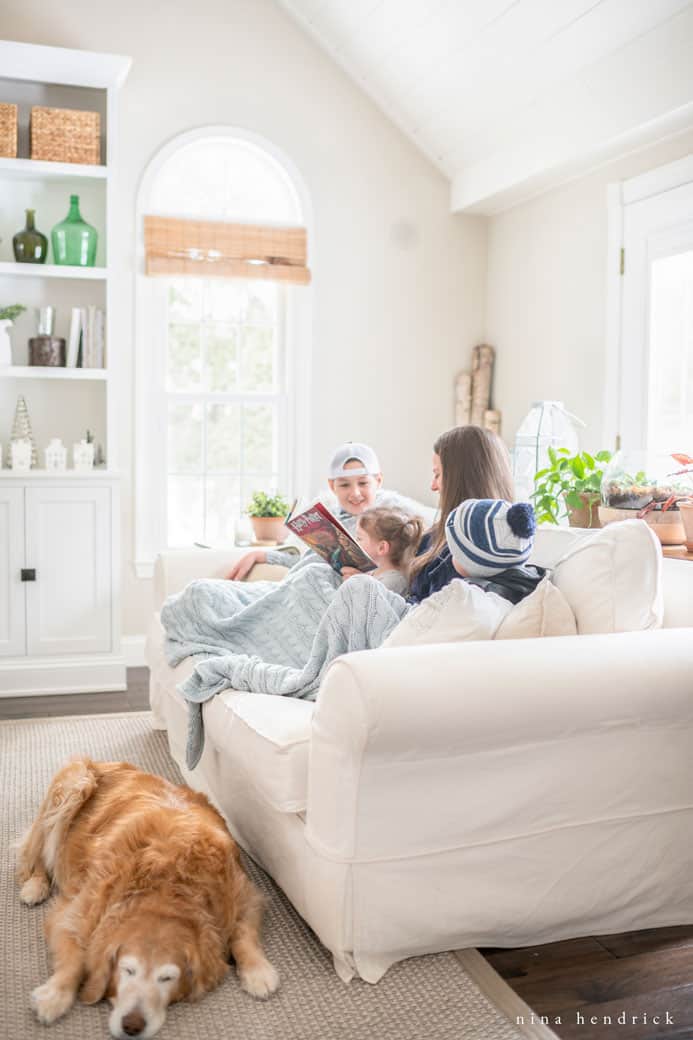 Family and dog cuddled up on a white slipcovered sofa reading a book with a blue blanket