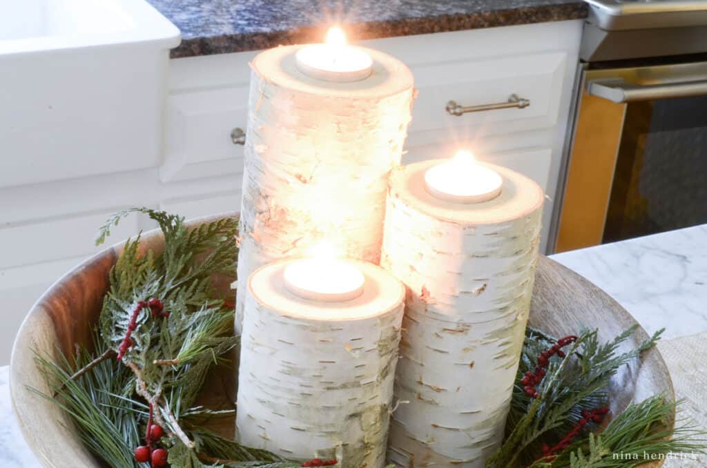 Three lit birch candles in a bowl with greenery.