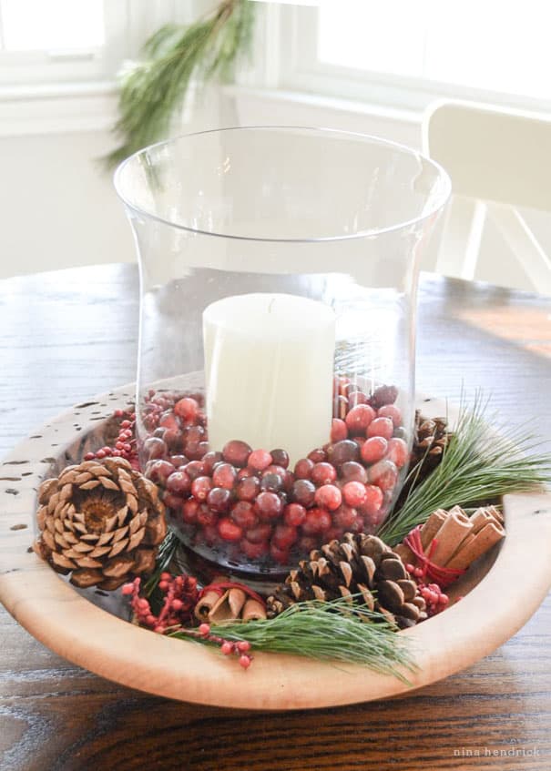 Cozy Christmas decorating ideas including this glass hurricane with cranberries in a wooden bowl surrounded by pine needles, pinecones, and cinnamon.