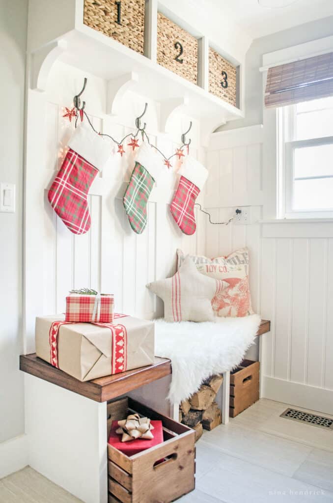 Mudroom bench decorated for a cozy Christmas with firewood, a faux fur throw, pillows, stockings, and lights