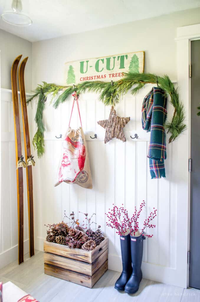 beadboard wall in a mudroom with boots, scarf, and vintage skis plus natural elements like pinecones and evergreens and a "U-Cut Christmas Trees" wooden sign