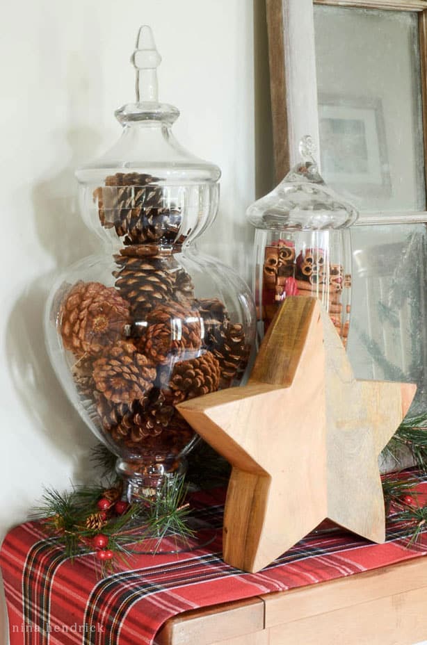 Christmas vignette decor featuring glass apothecary jars filled with pinecones and cinnamon sticks with a wooden star.