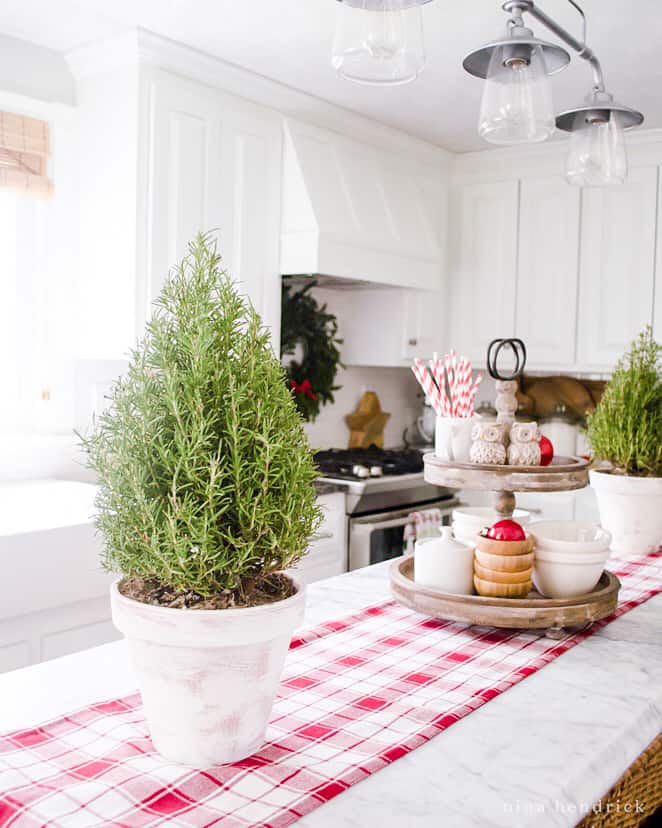 Easy Christmas Decorating ideas in the kitchen — a decorative tiered tray and rosemary topiaries in white pots with a red plaid runner. 
