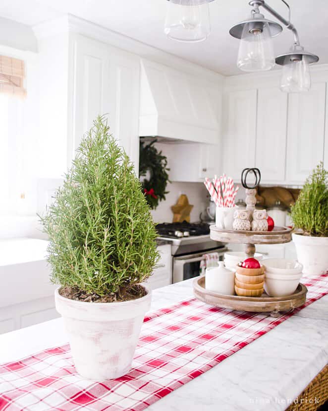Easy Christmas Decorating ideas in the kitchen — a decorative tiered tray and rosemary topiaries in white pots with a red plaid runner.