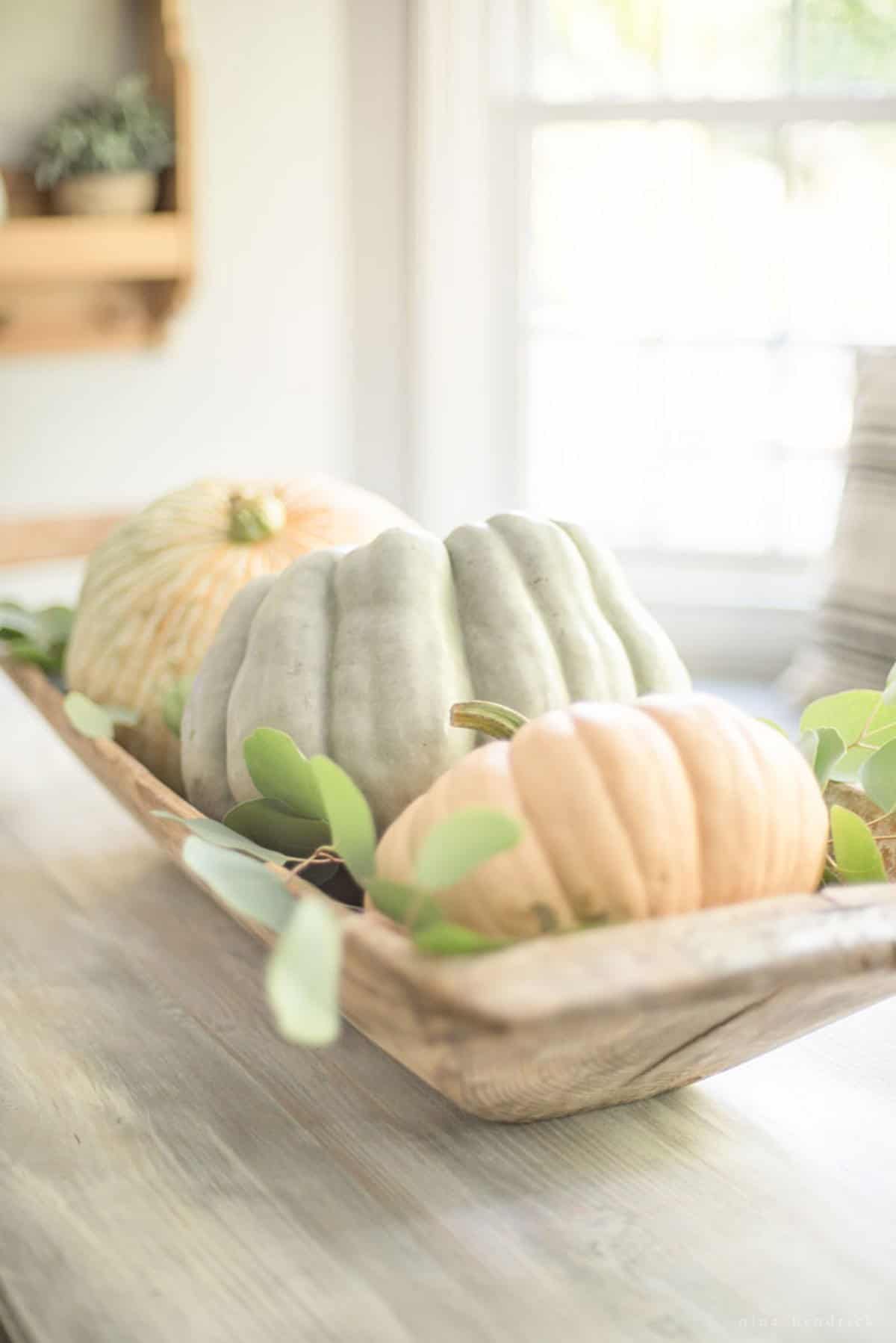 A wooden bowl filled with pumpkins and eucalyptus leaves.