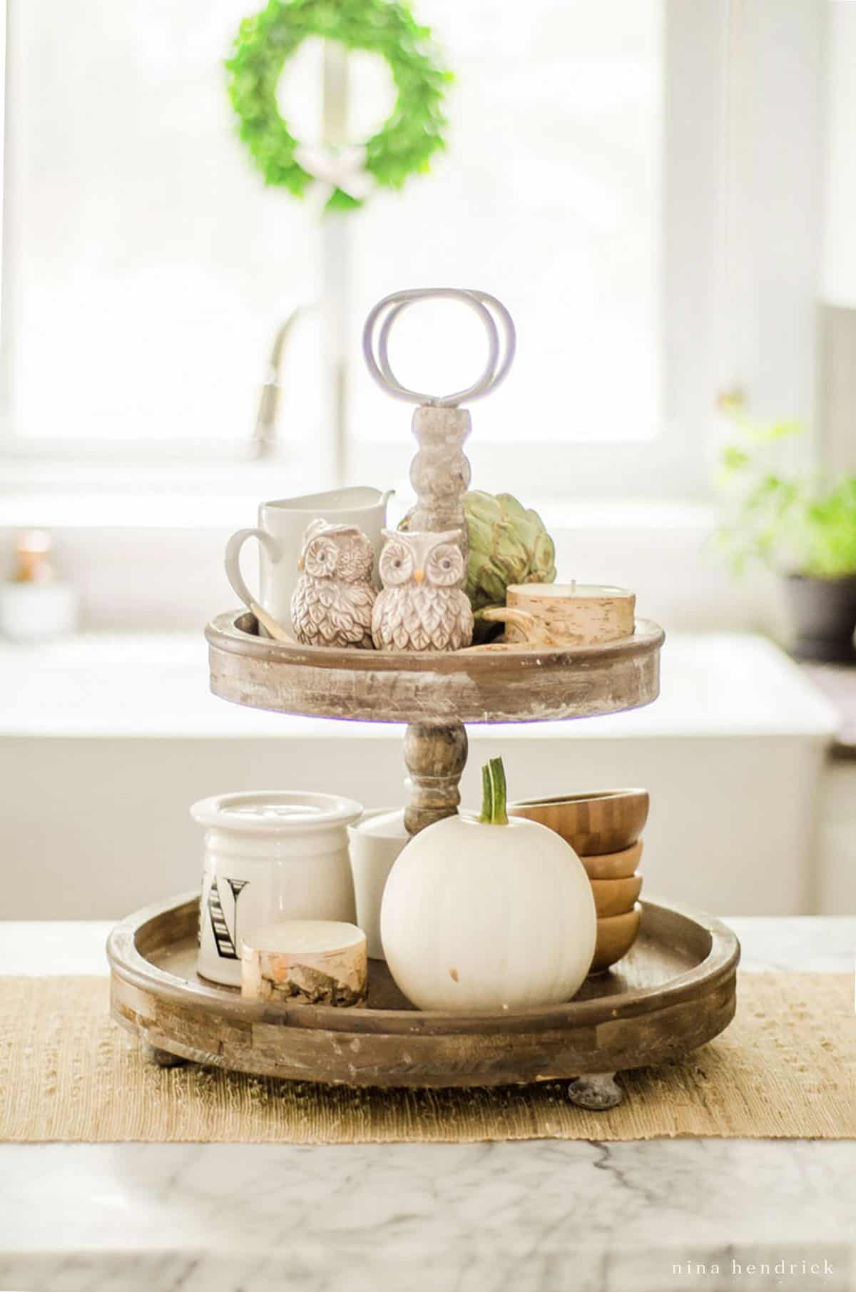 Three tier tray fall centerpiece in the kitchen with pumpkins and a wreath on it.