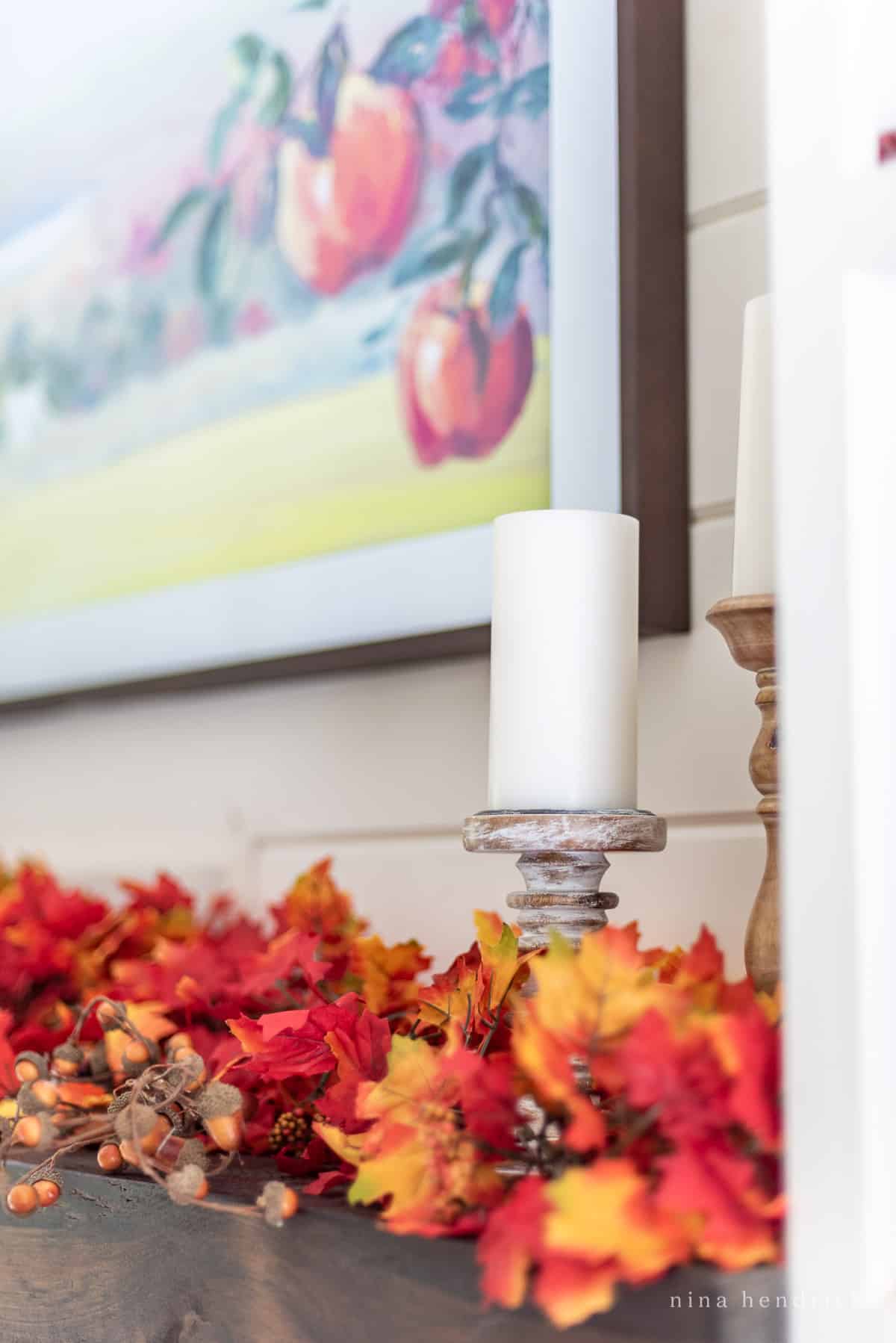 Close-up of a white pillar candle on a candle holder with fall leaves garland on a mantel.