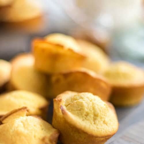 Close-up of several golden-brown sweet cornbread muffins stacked and spread out on a wooden surface, with soft focus in the background.