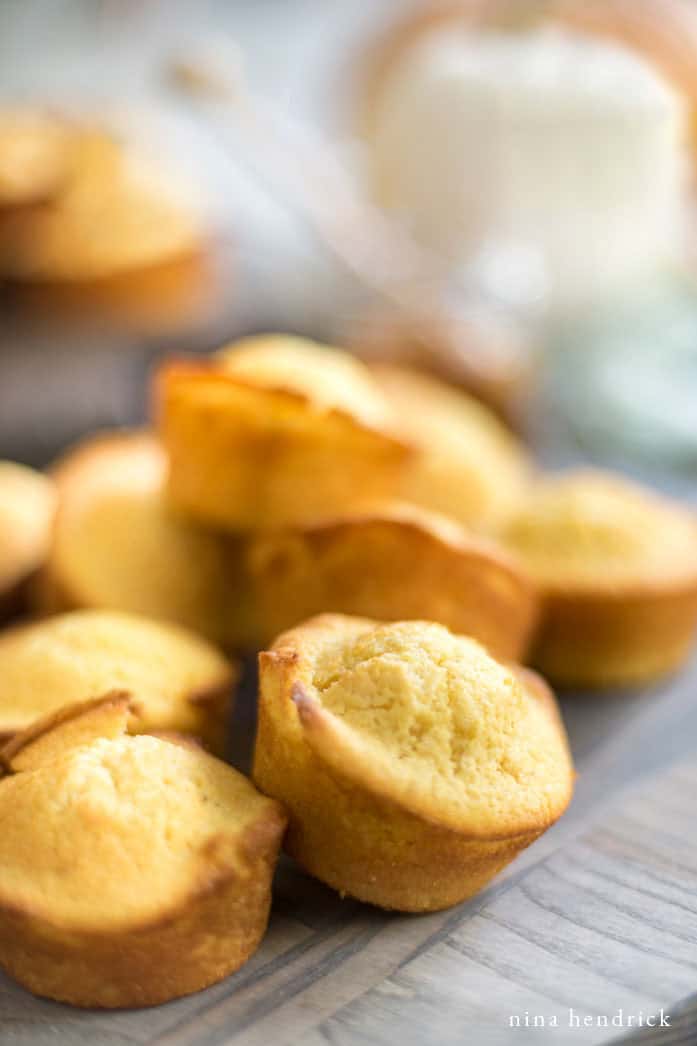 Close-up of several golden-brown sweet cornbread muffins stacked and spread out on a wooden surface, with soft focus in the background.