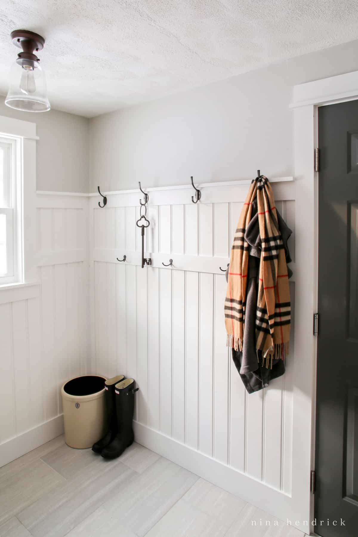 Home Organization ideas in the entryway. Beadboard wall with hooks for hanging coats. Crock for holding umbrellas. Boots on a tile floor.