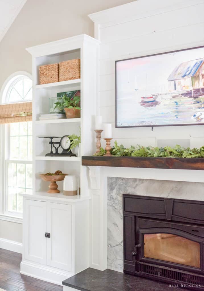 Built-in bookcase with decor and a mantel covered in greenery with a Frame TV above it featuring coastal artwork