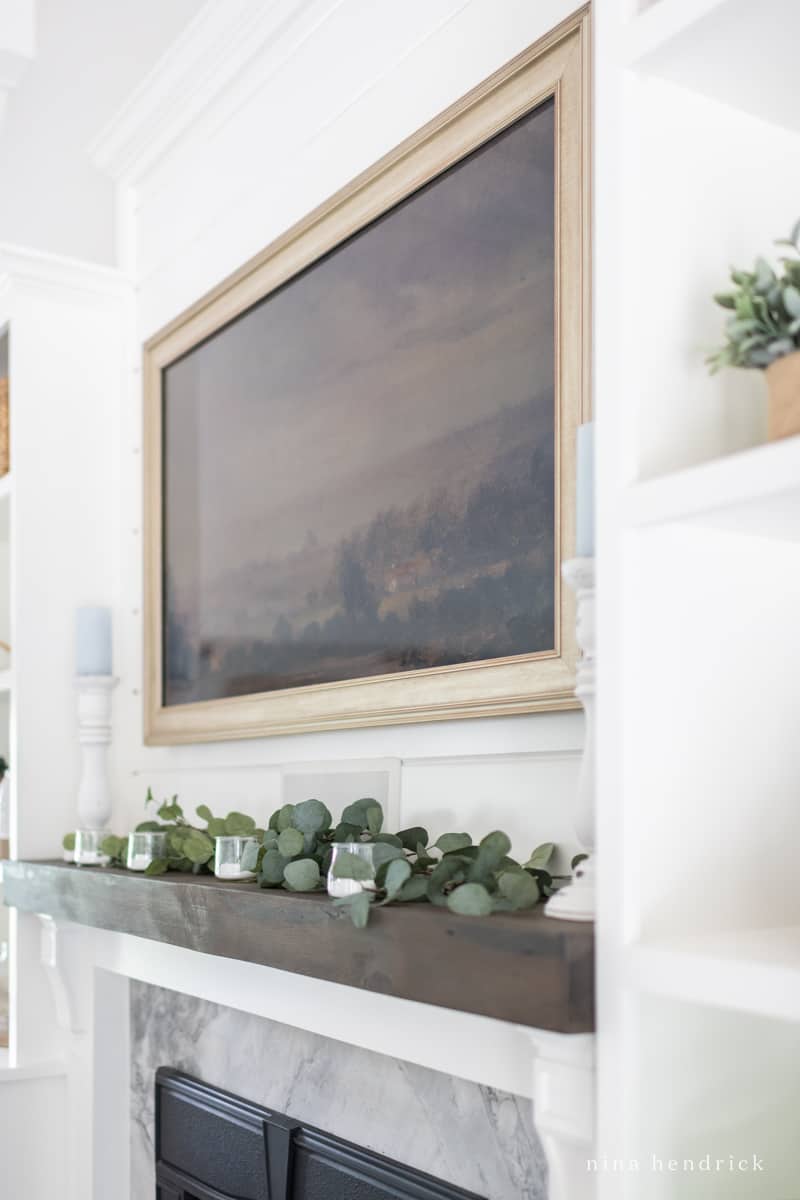 Dark wood mantel with greenery and a TV with gold frame and artwork