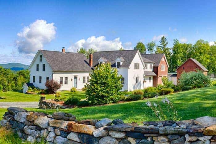 A New England farmhouse in the middle of a grassy field with a stone wall