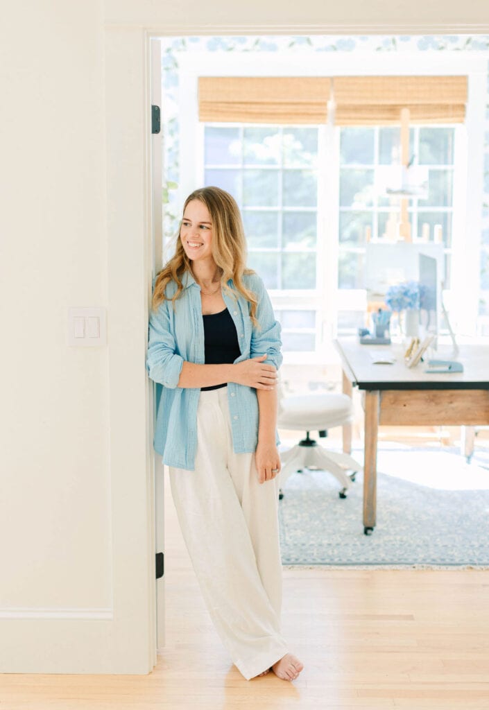 A woman stands barefoot in a doorway, wearing a light blue shirt over a black top and white pants, with a bright office space visible in the background.