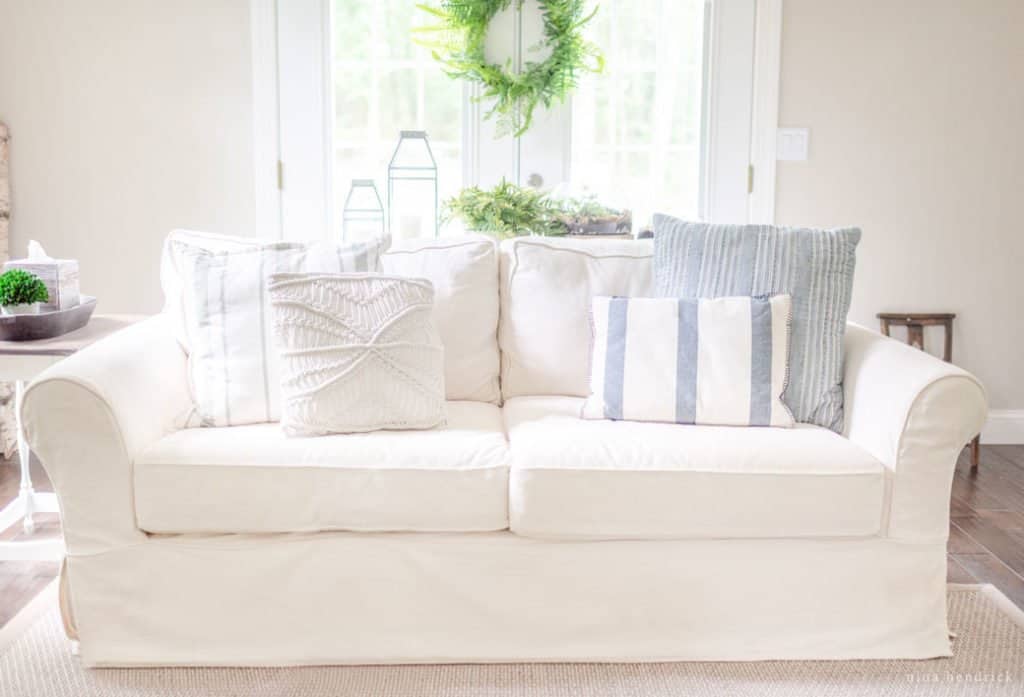 White couch with neutral throw pillows in a bight and sunny living room with a fern wreath.