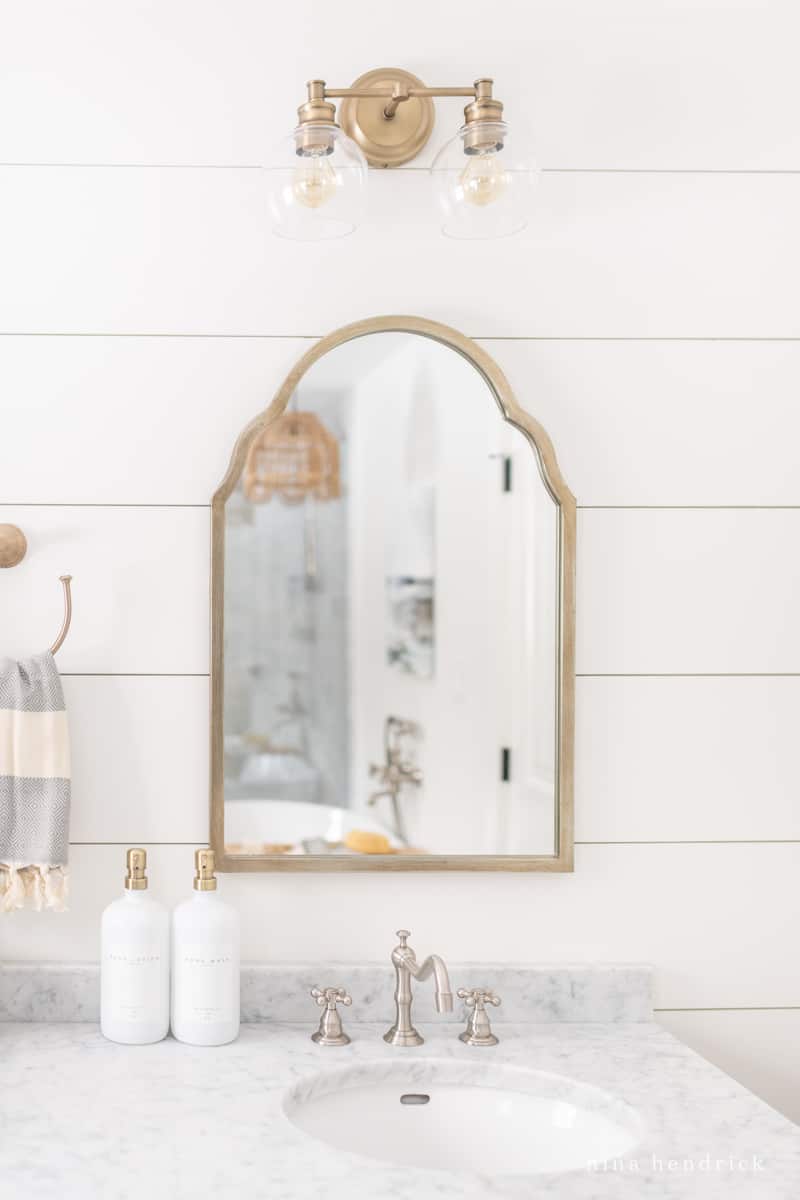 gold bathroom mirror with plank wall and sconce above sink