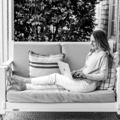 Nina Hendrick reclines on an outdoor couch while using a laptop. She is barefoot, dressed in casual attire, and surrounded by pillows. The setting appears relaxed with greenery visible in the background.