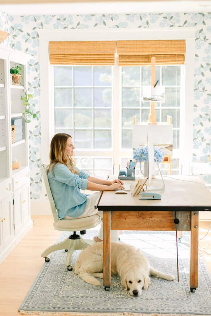 Nina Hendrick works on a computer in a bright room with floral wallpaper. She sits on an office chair at a desk while her dog lies comfortably on the floor beneath her.