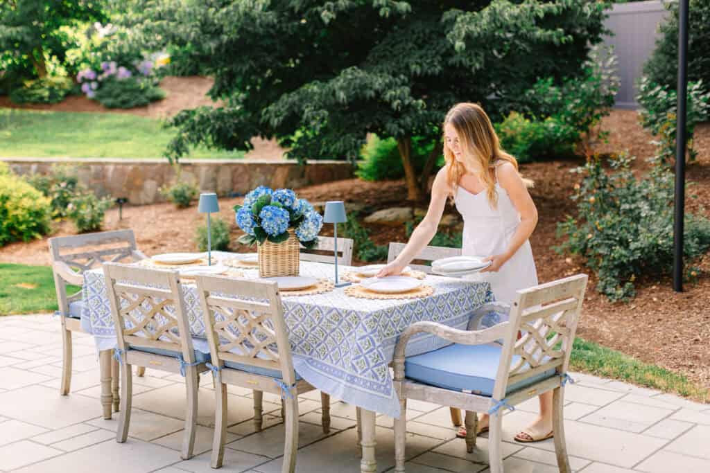 Nina Hendrick effortlessly sets a dining table with blue chairs and a floral centerpiece on a patio surrounded by greenery, all while wearing a white dress.