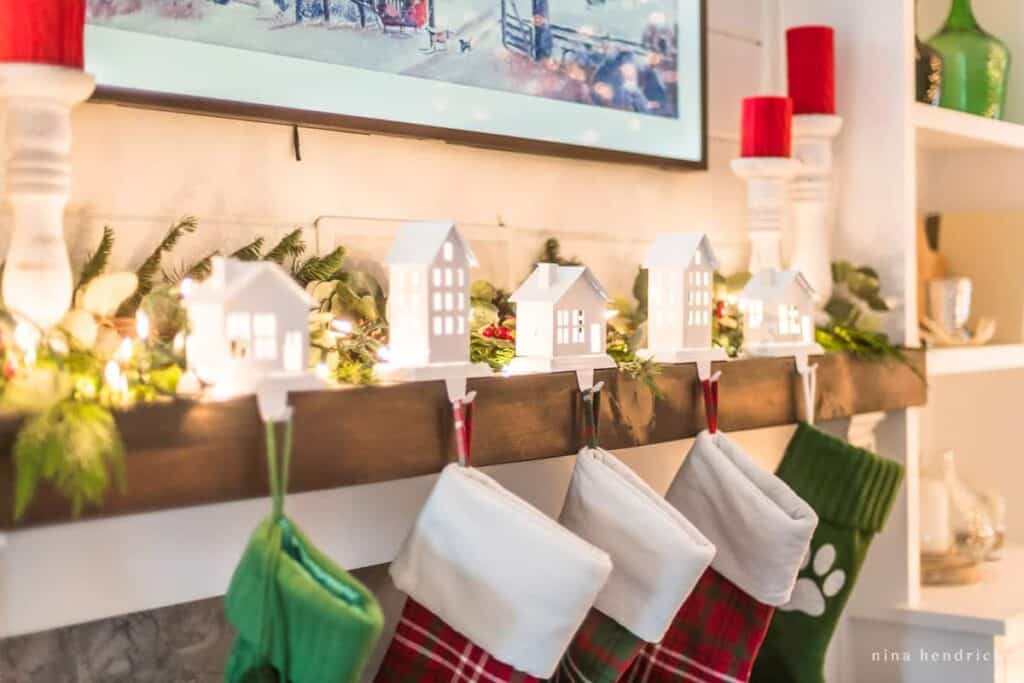 Christmas mantel decor with stockings, village themed stocking holders and a Frame TV with winter art
