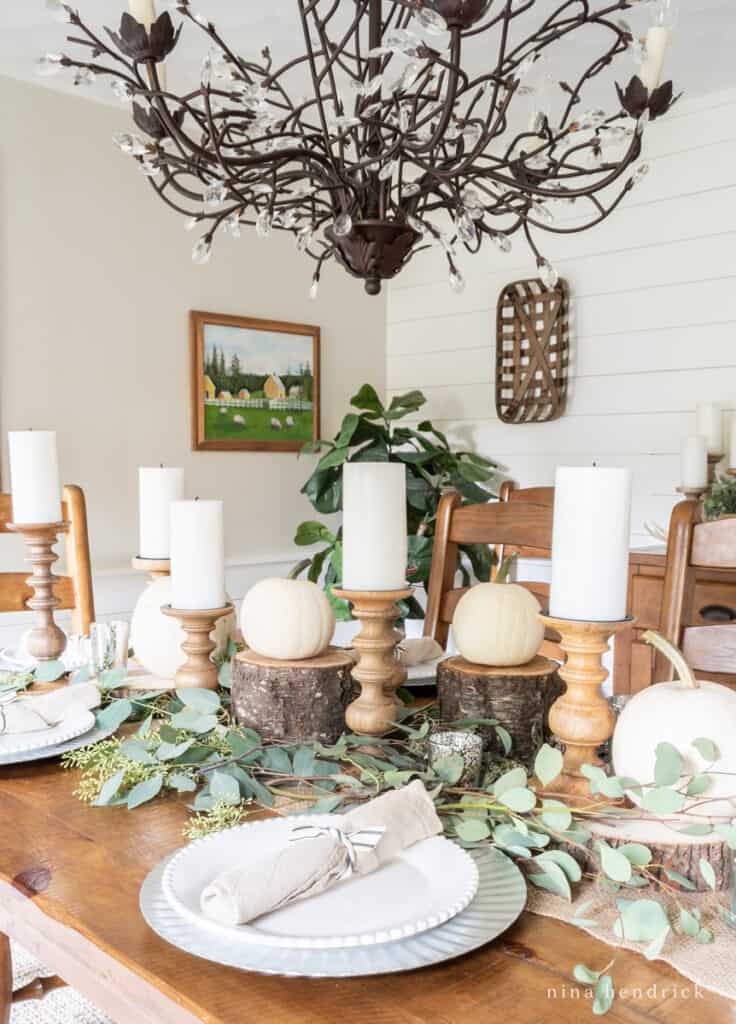 Neutral and rustic Thanksgiving centerpiece with wooden candle holders, wood slices, white pumpkins, and seeded eucalyptus.