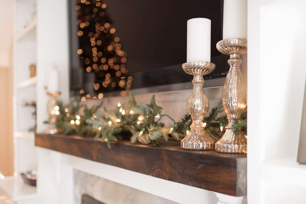 Simple mantel Christms decor — an evergreen garland, string lights, a mercury glass candle holder and white candle