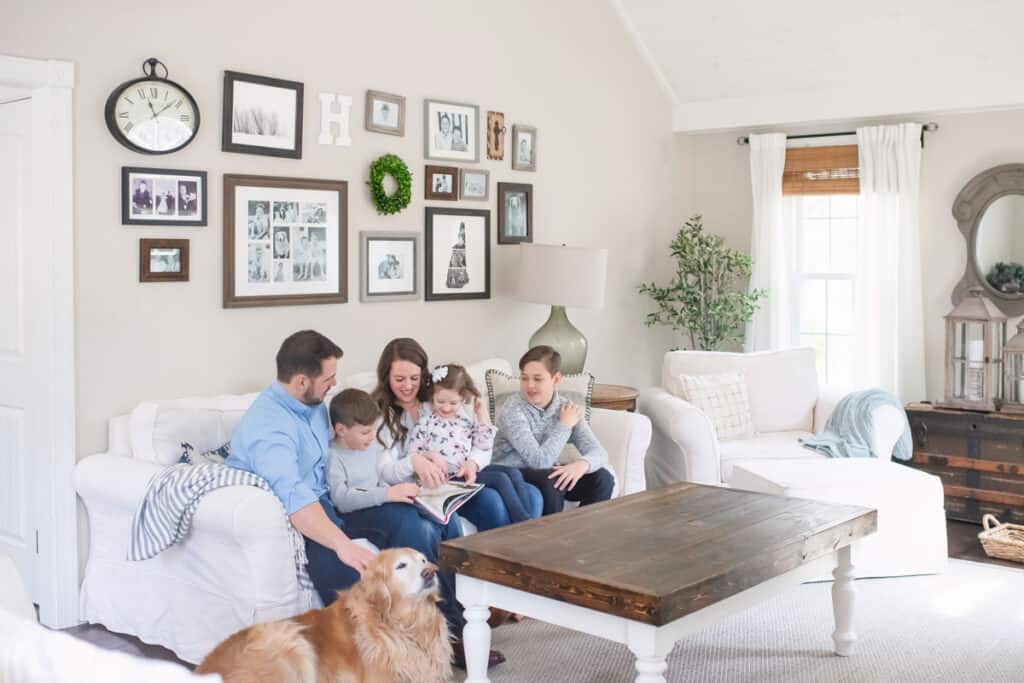 Family sitting on a white couch reading a book with a golden retriever and a gallery wall.