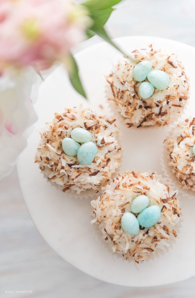 Four bird nest cupcakes topped with toasted coconut and small blue speckled candies, arranged on a white plate.