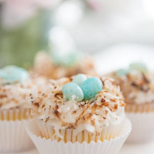 Bird nest cupcakes topped with toasted coconut and blue speckled candy eggs in white wrappers.