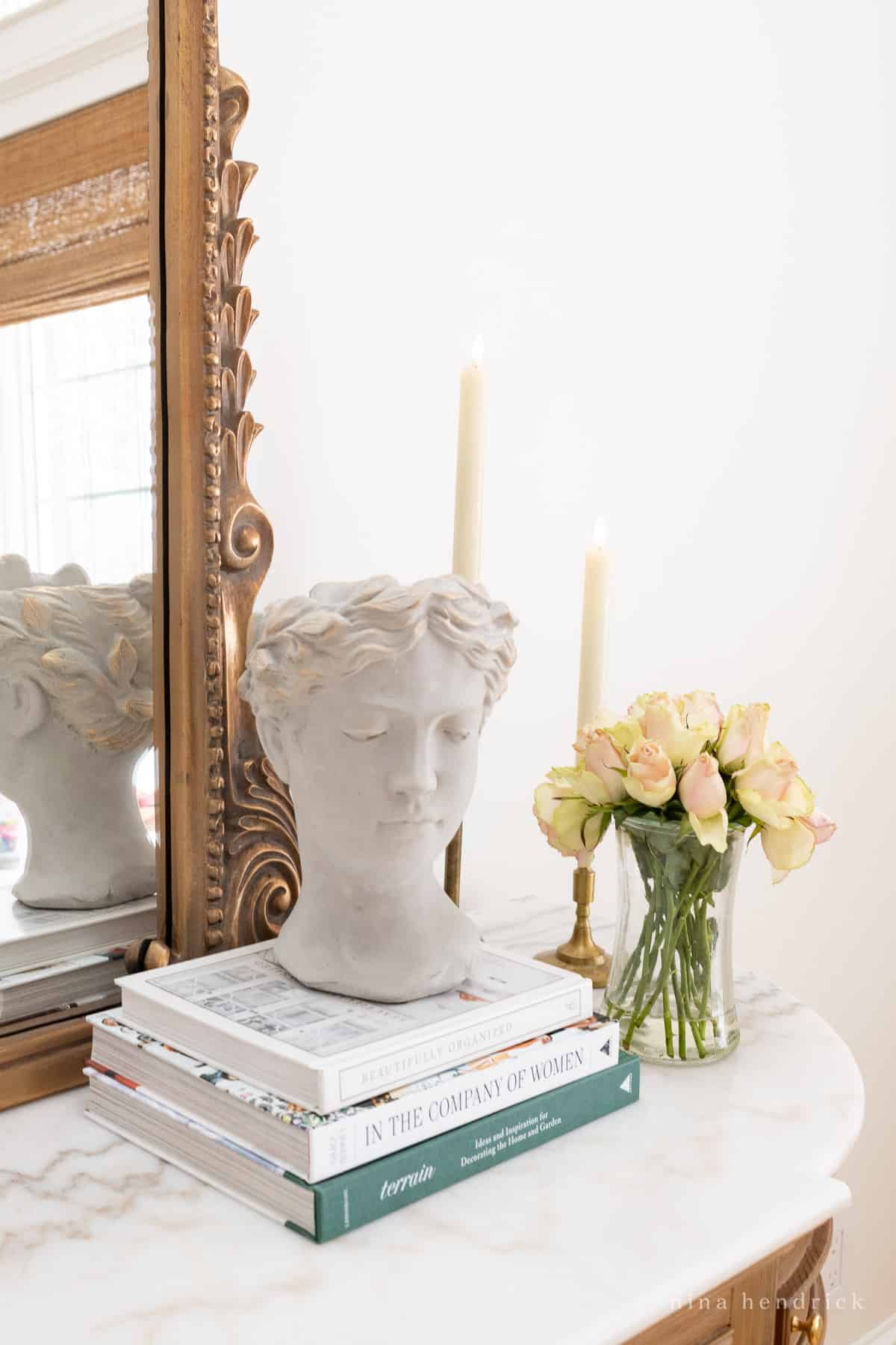Vignette decor with a stack of books, roman bust, a vase of flowers, and candle sticks