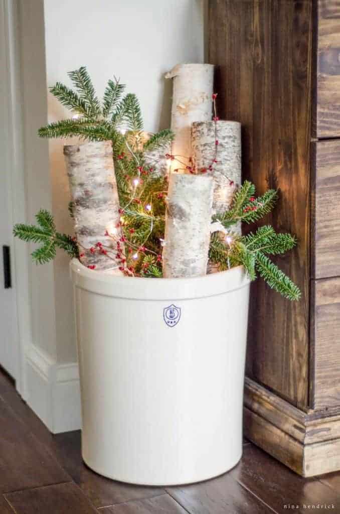 Christmas arrangement in a 5-gallon crock with birch logs, greenery, red berries, and string lights.