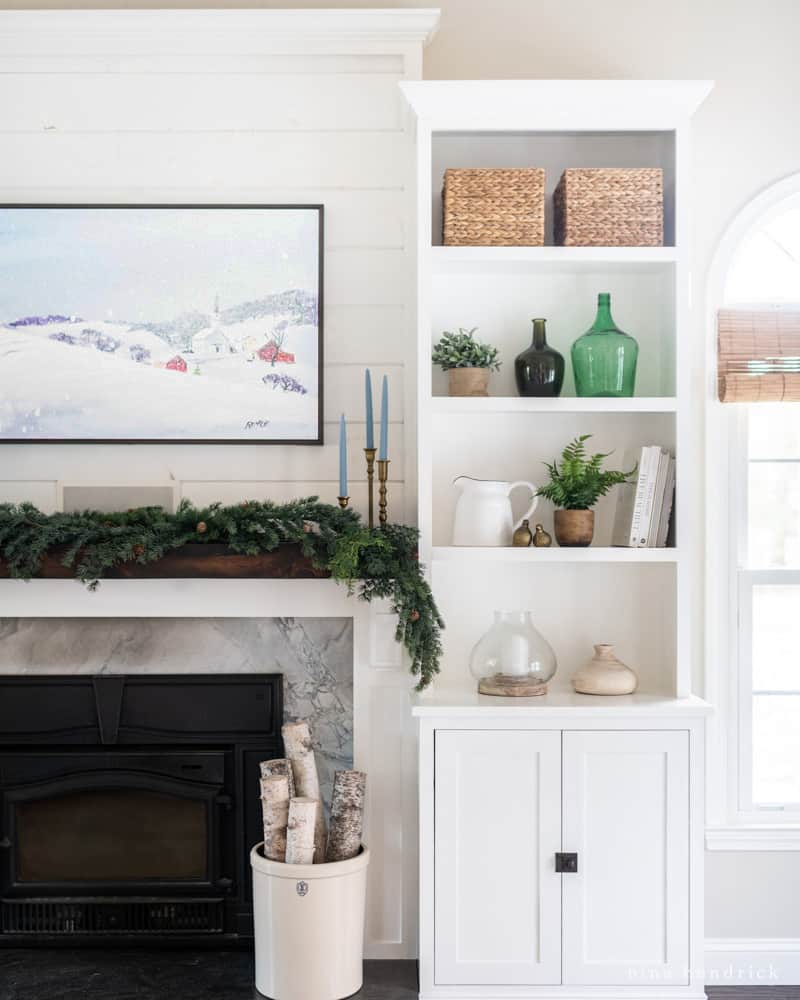 Mantel decorated for winter with evergren garland and candlesticks with a winter painting and birch logs.